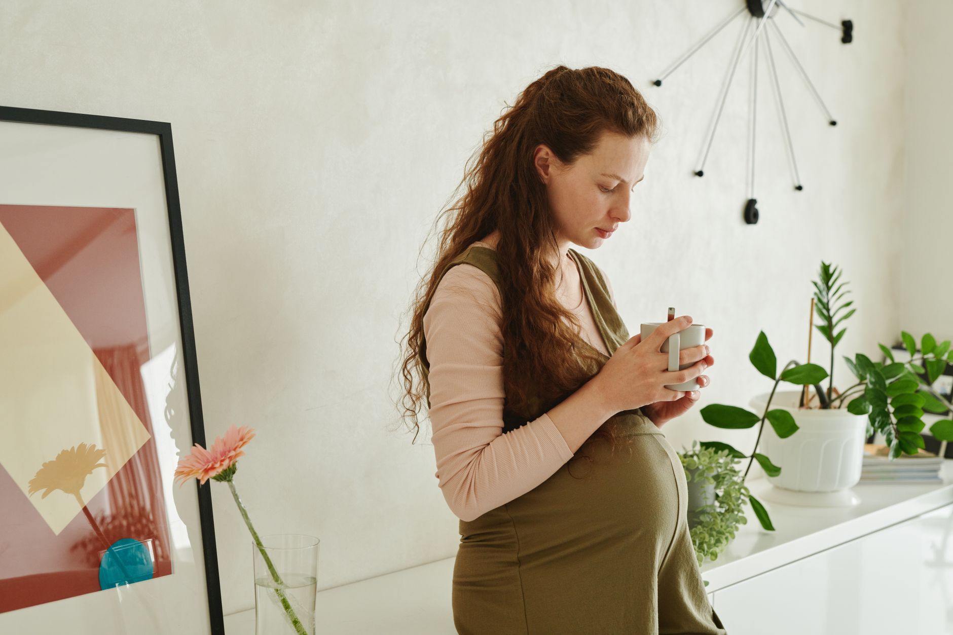 Pregnant woman holding a cup of ginger tea by a window, contemplative expression